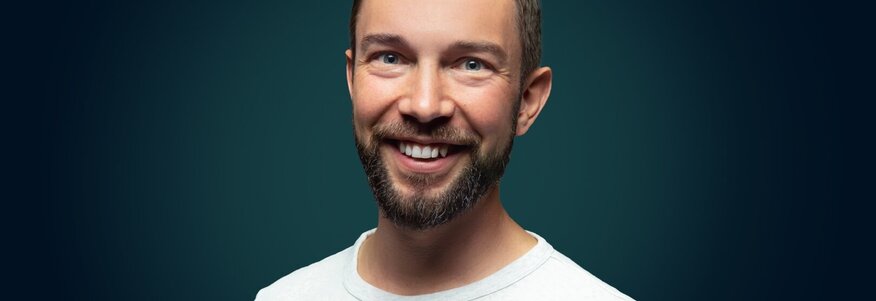Hier sehen Sie einen Mann mit braunem Haar und braunem Bart, der ein weißes T-shirt trägt und lächelt. 